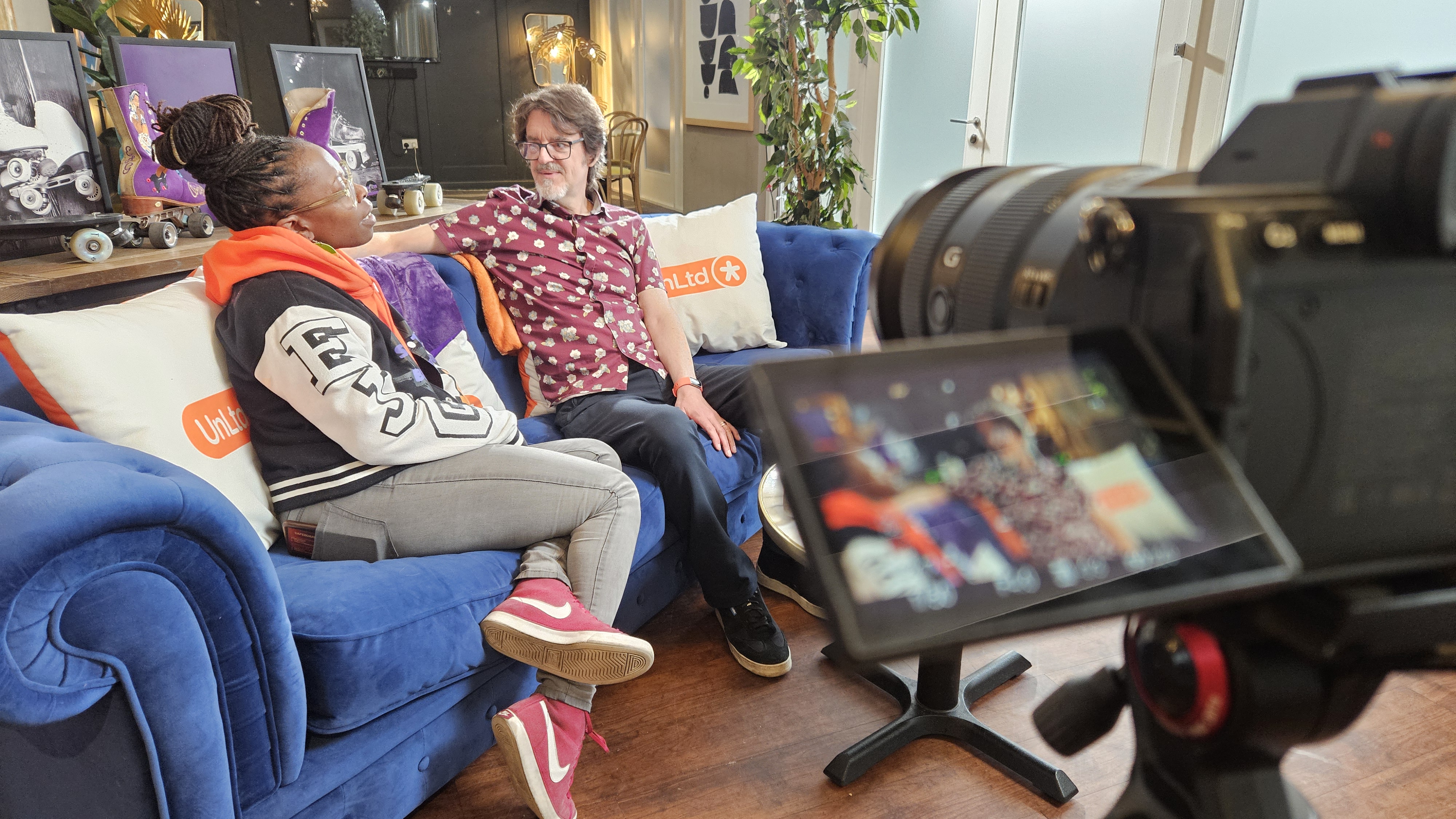 Empress and John sit on a blue sofa together while we look through a camera that is filming them.