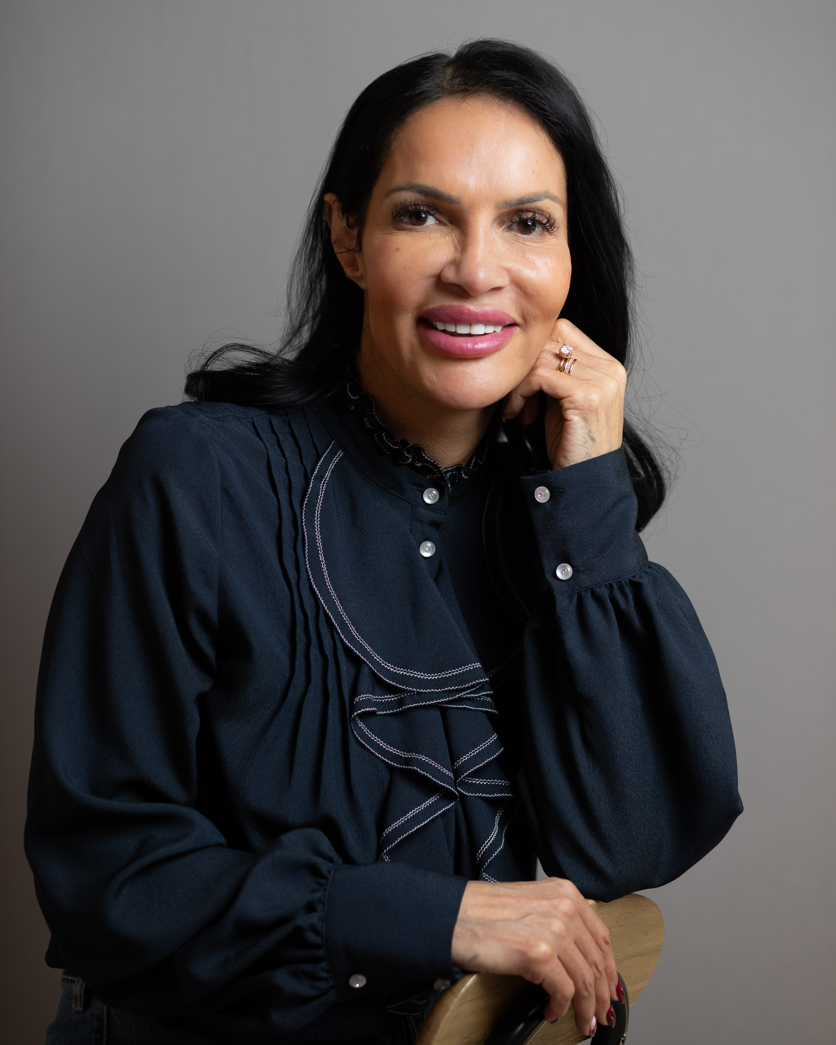 Image shows Lilly smiling at the camera with her face rested on one hand against a grey muted background.