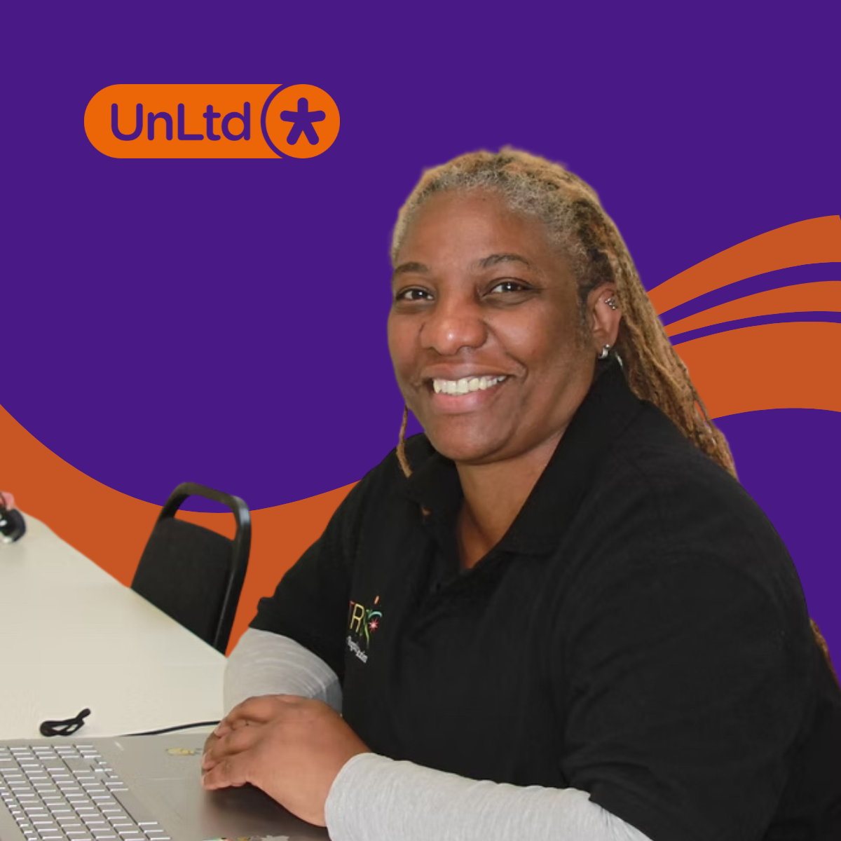 Image shows Jackie Brooks smiling at the camera whilst working on a laptop at a desk with a purple background and an orange wave behind Jackie.