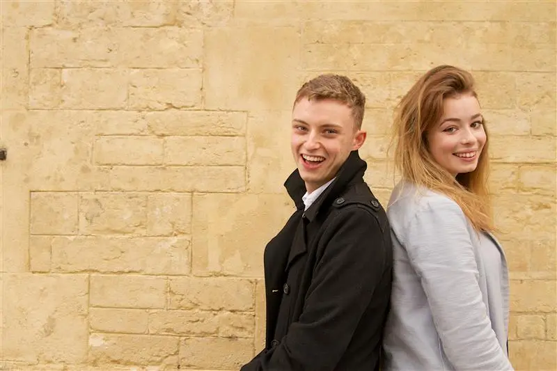 A young man and woman stand back-to-back, both smiling at the camera. The man wears a black coat over a white shirt, while the woman wears a light blue jacket. They are positioned against a textured beige stone wall, giving the image a warm and natural background.