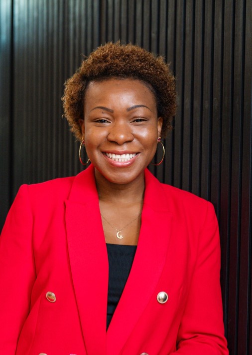 Success stands facing the camera, smiling, with a red suit and gold necklace and hoops.