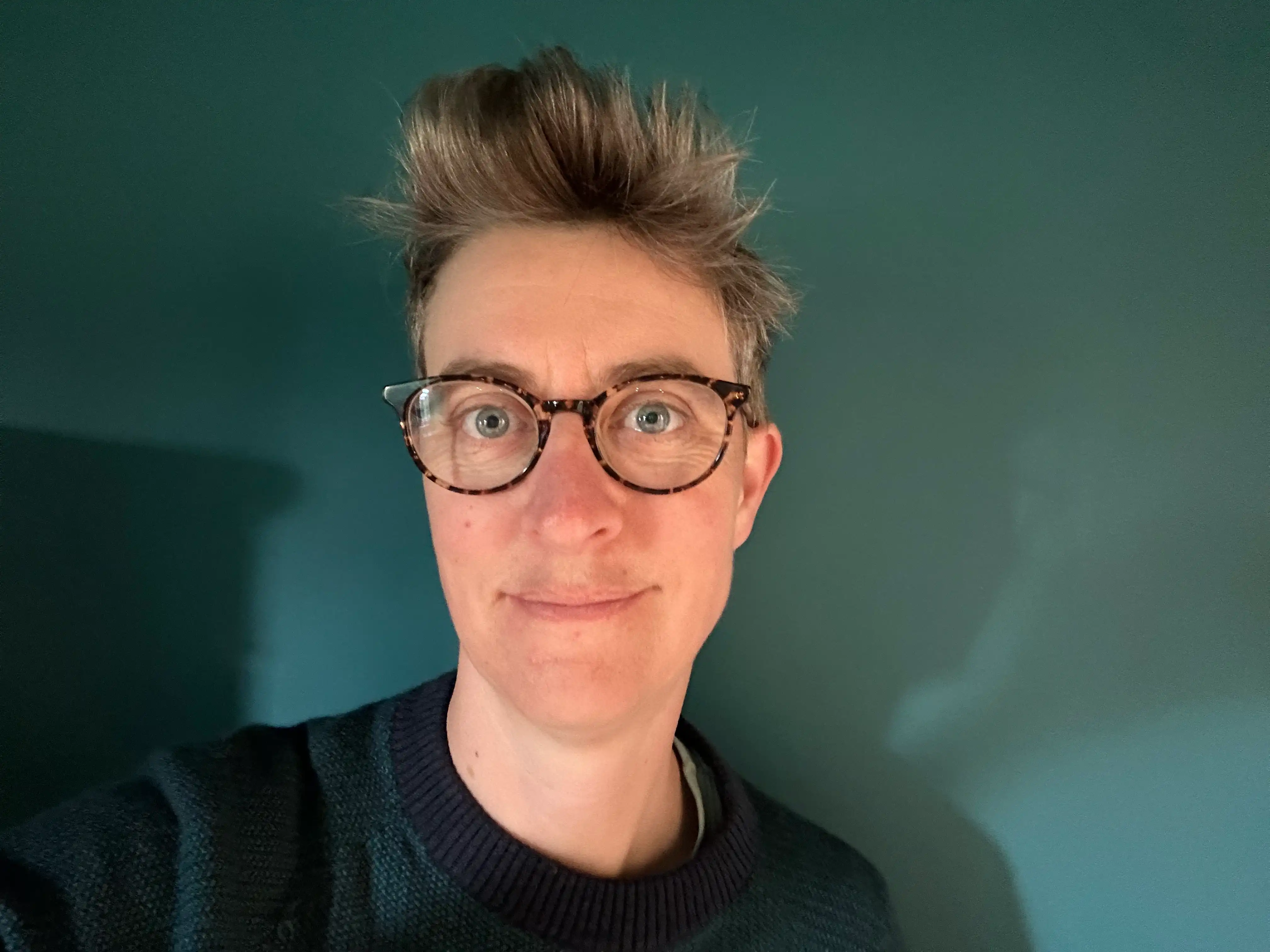Jill Corbyn, founder of Neurodiverse Connection CIC, with short hair and tortoiseshell glasses, is wearing a dark green jumper with a light green background and is smiling at the camera.