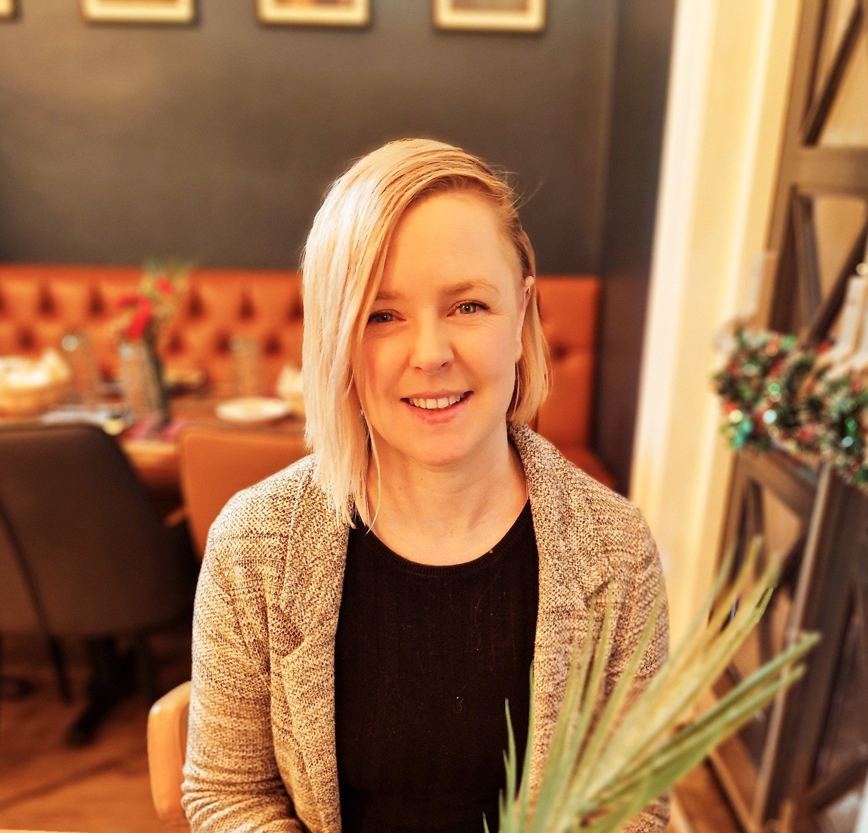 Image shows Aimee, with blonde hair swept over one side, in a black t-shirt and a grey cardigan, in the background you can see the leather brown chairs of a restaurant.
